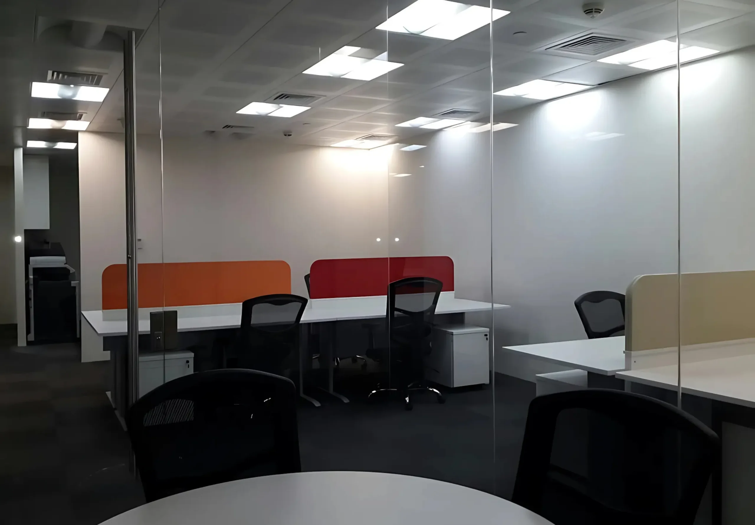 Modern office space with glass partitions, individual workstations, and overhead lighting.