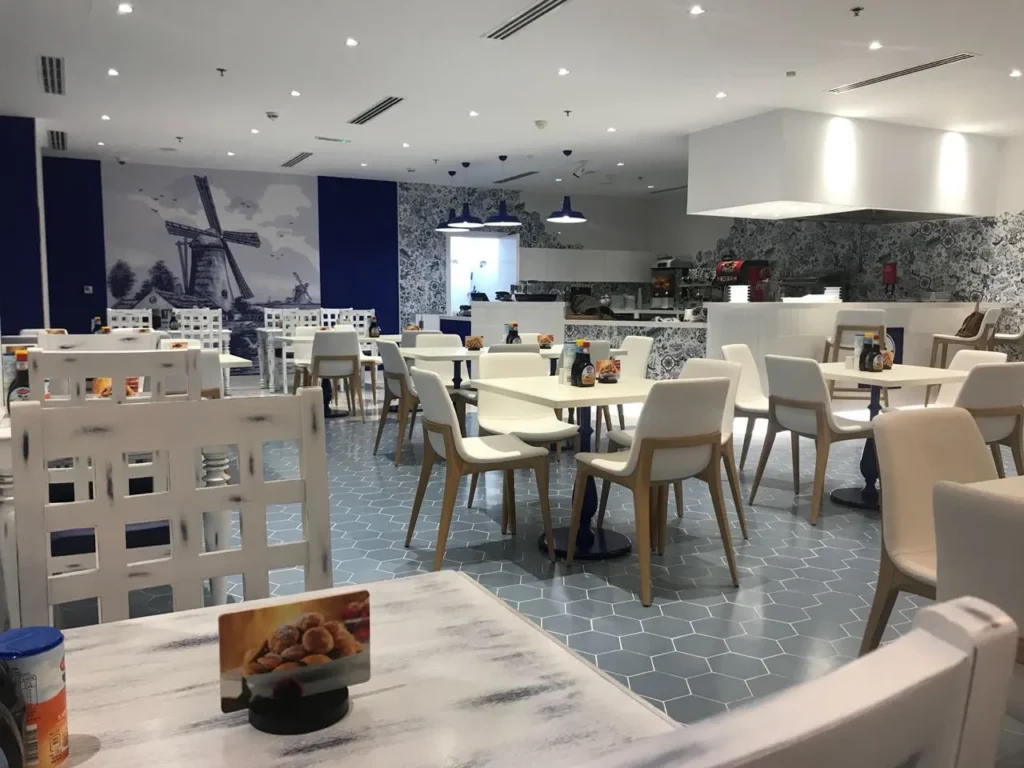 Bright café interior with Dutch-inspired design, featuring round tables, white chairs, and windmill wall art.