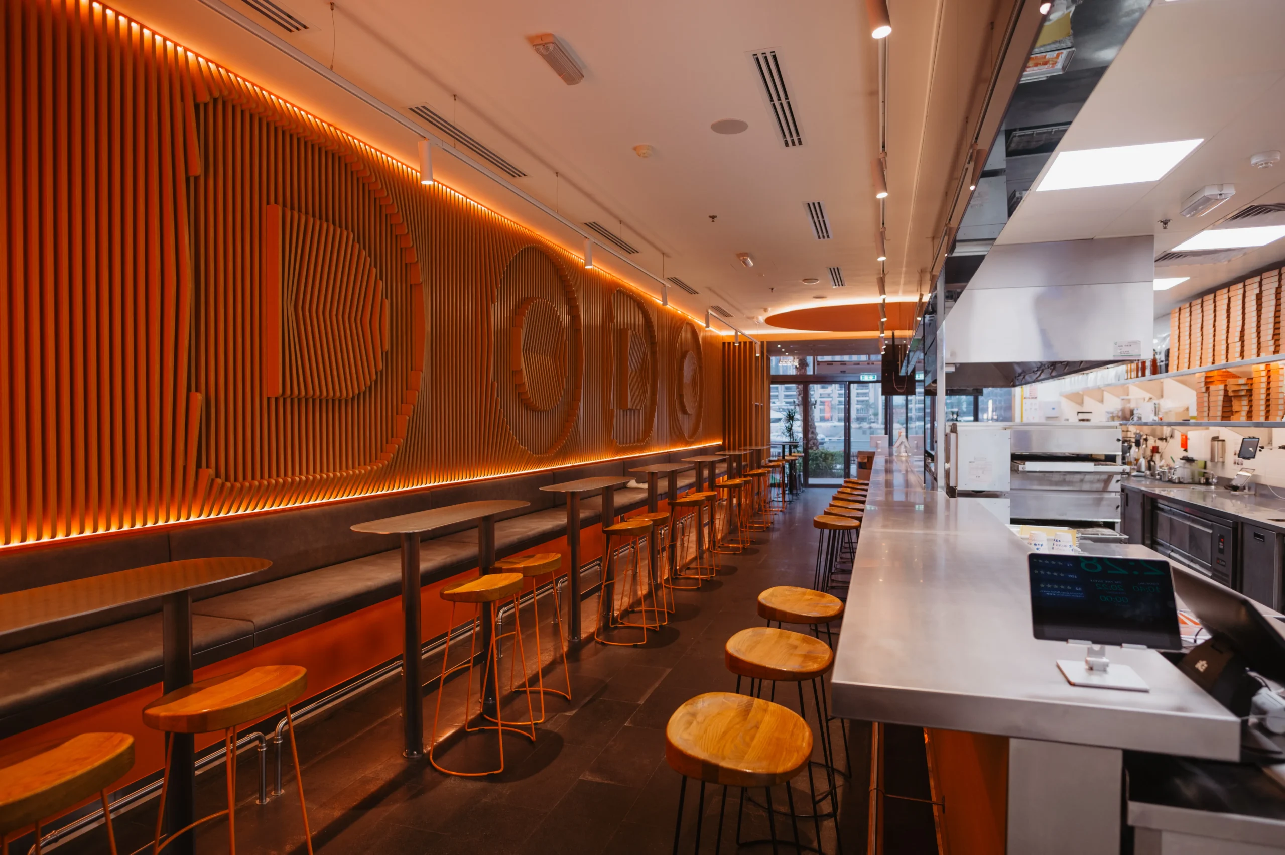 Trendy pizza restaurant interior with orange accent lighting, wood seating, and an open kitchen layout.